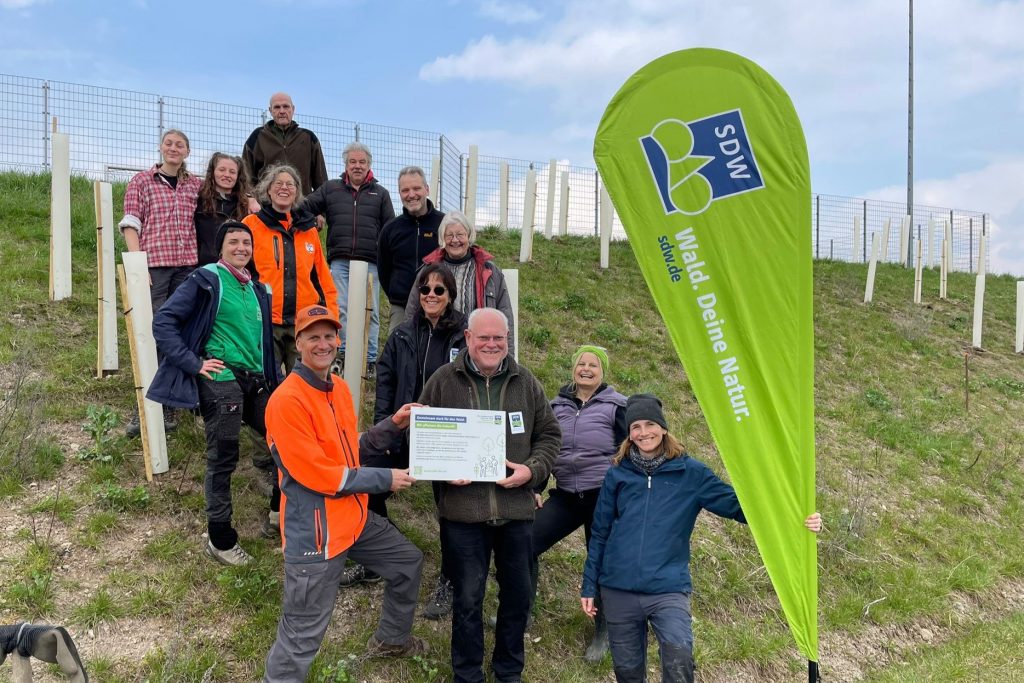 Eine Gruppe von Menschen steht an einem frisch bepflanzten Hang. Im Vordergrund steht eine BEachflag mit der Aufschrift "SDW BW". Zwei Männer halten ein Schild in der Hand.